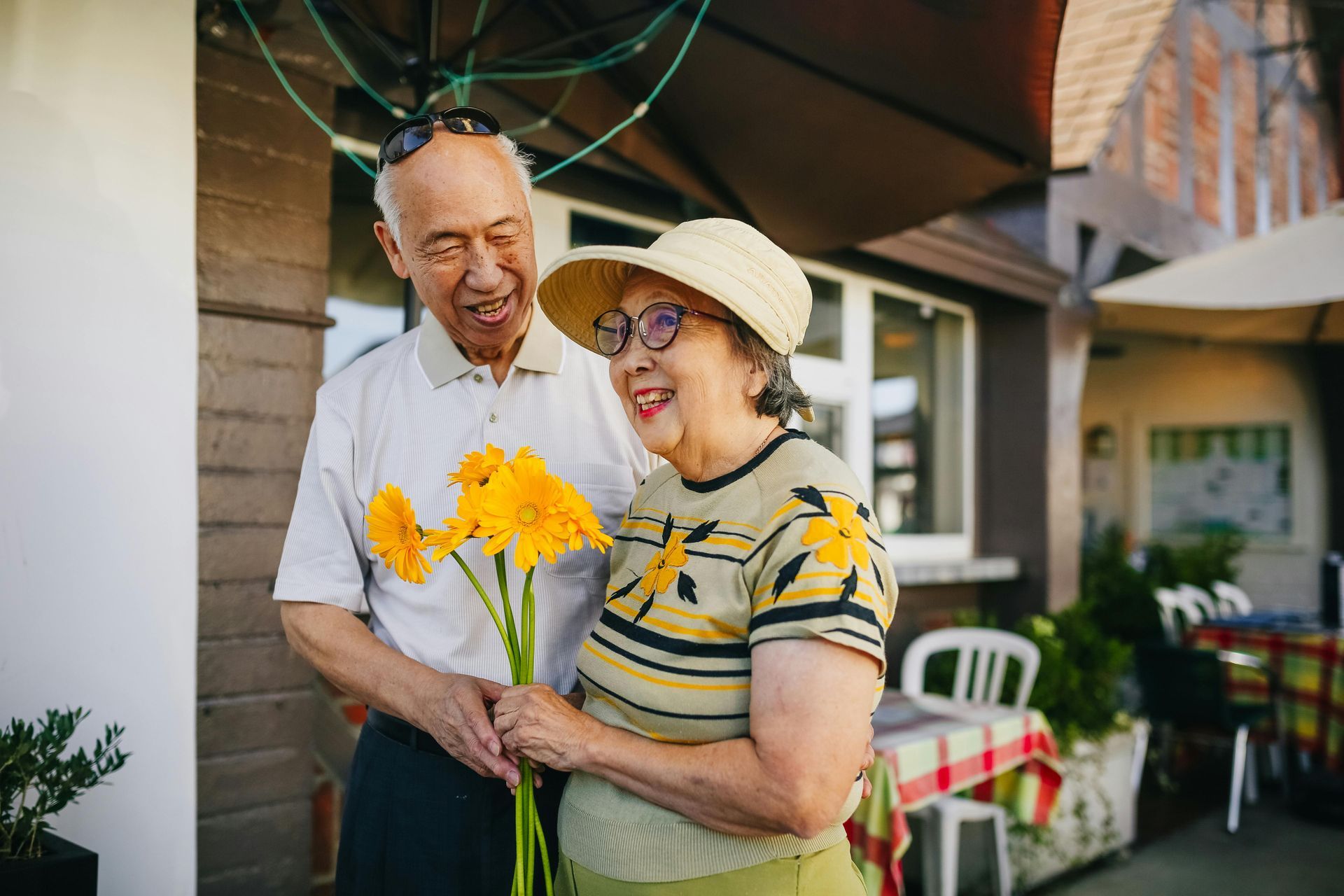 A person offers a bouquet of yellow flowers to another person outdoors, both smiling warmly in a casual patio setting at Lexington at Champions in Houston, TX..