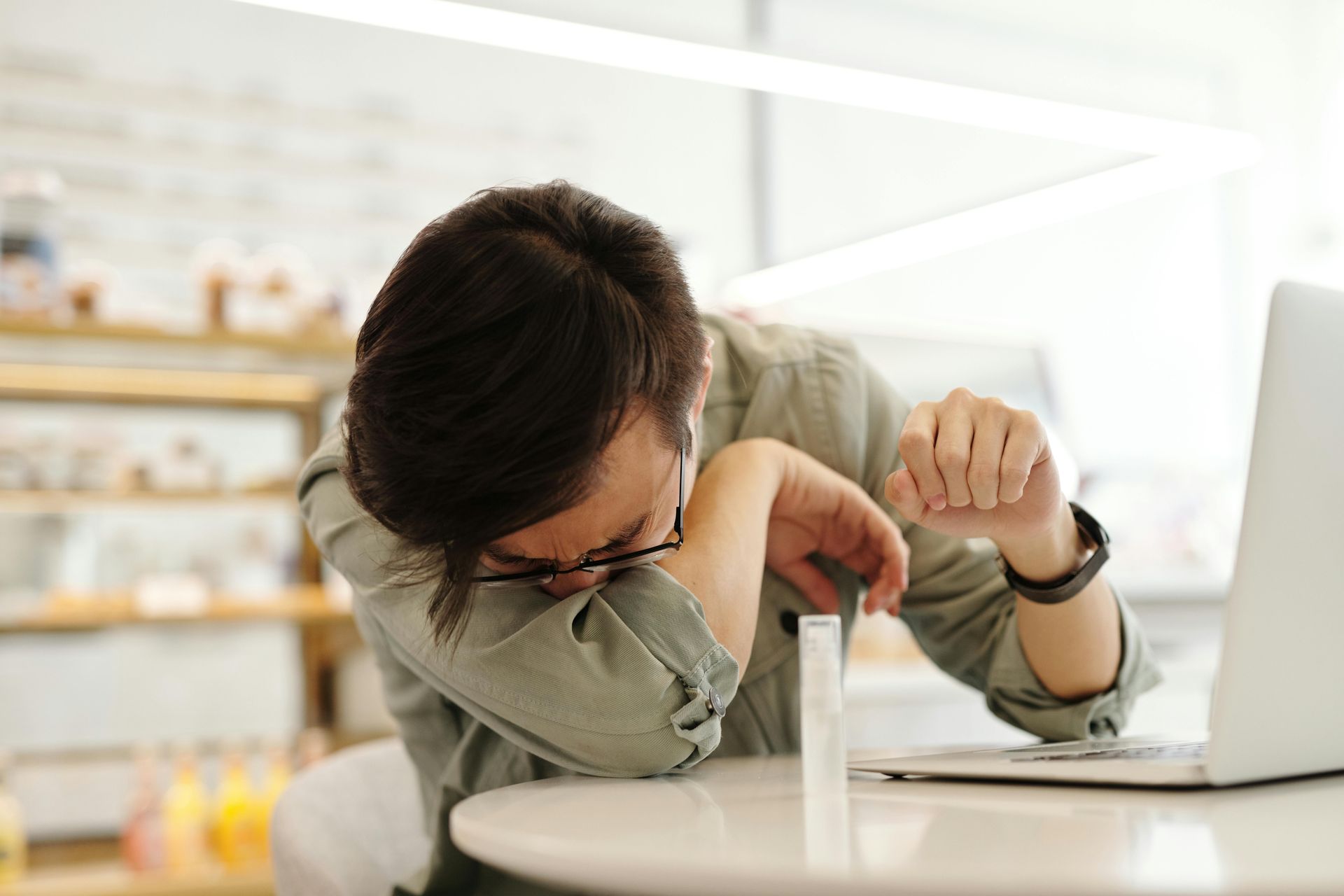 A man is sitting at a table with a laptop and rubbing his eyes.