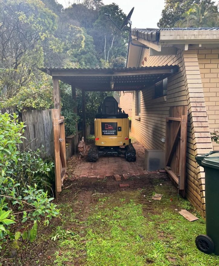 A Yellow Excavator Is Parked in A Driveway Next to A Brick Building — Norcoast Diggers in Ballina, NSW