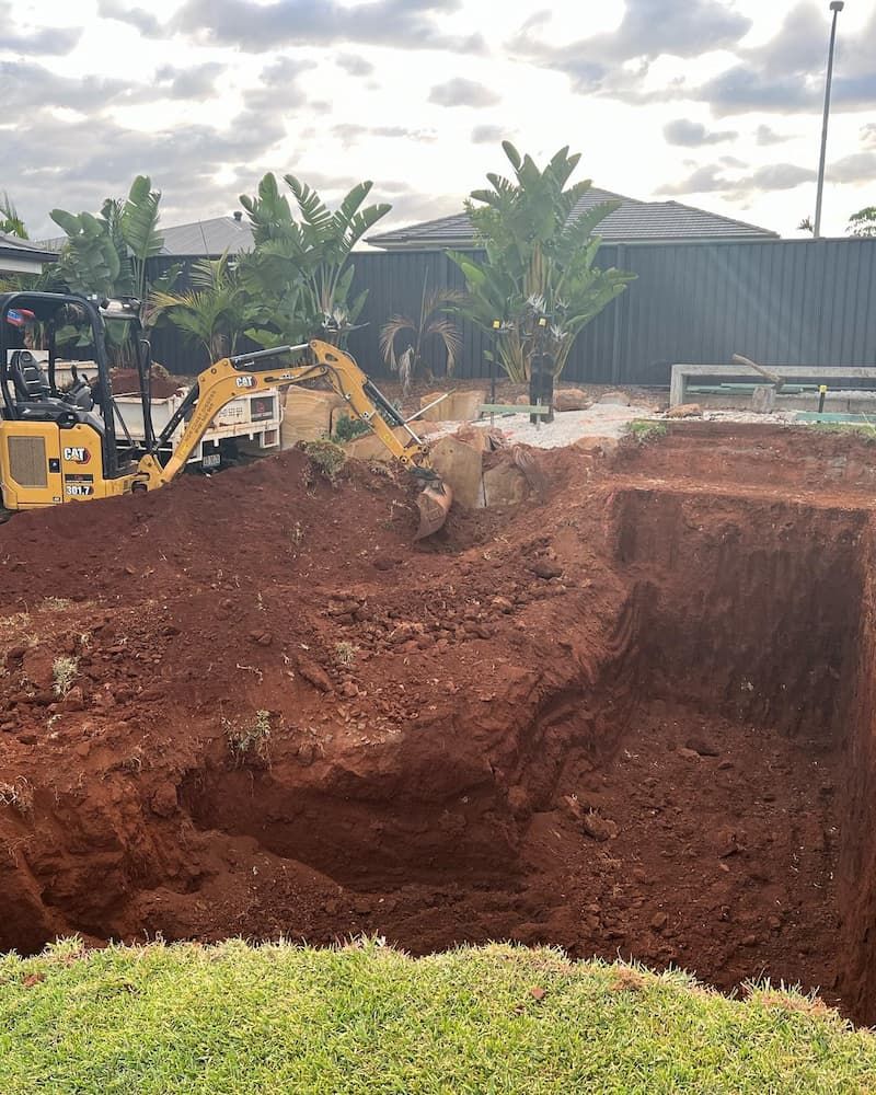 A Yellow Excavator is Digging a Hole in the Dirt in Front of a House — Norcoast Diggers in Byron Bay, NSW
