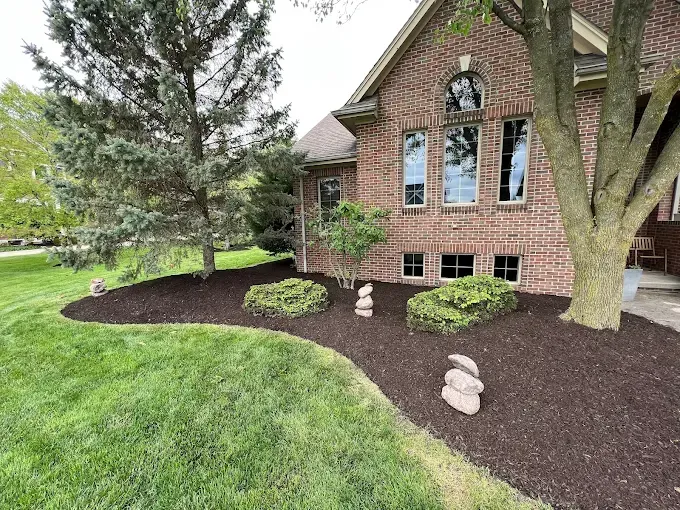 A brick house with a mulched flower bed, trimmed bushes, and a tree. Green lawn in front.