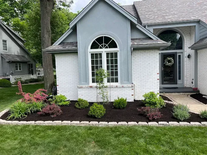 House with a landscaped front yard; white brick, blue-gray trim, and a black door. Mulch and varied shrubs.