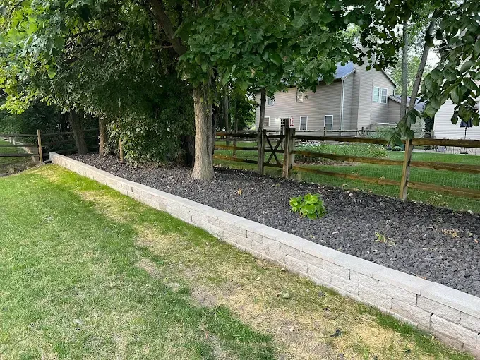 A low retaining wall bordering grass and mulch-covered planting area, with trees and a fence in the background.