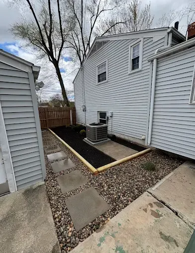 Backyard with pathway, flower beds, and a building with air conditioning unit.