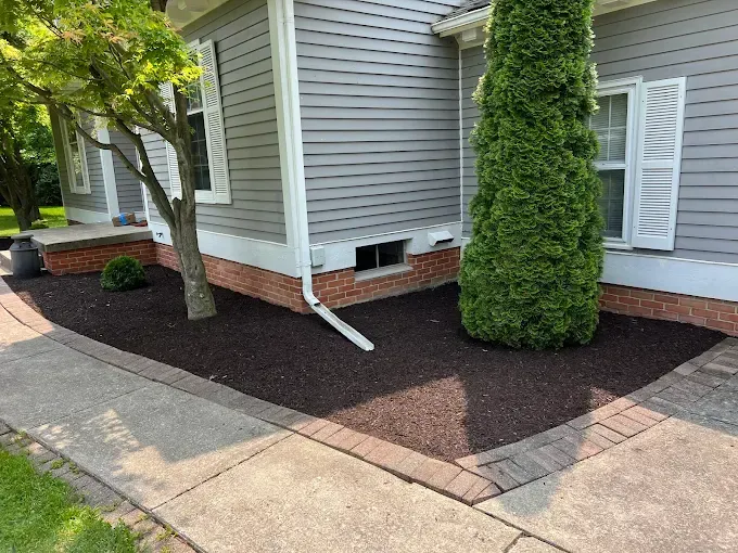Side of a house with gray siding, brick foundation, and dark mulch landscaping.