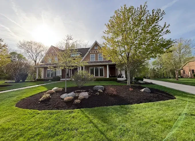 Two-story brown house with a porch, landscaped yard, large trees, and a sunny sky.
