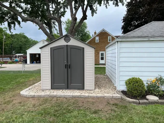 A white and gray storage shed in a backyard, near a white garage and a yellow house.