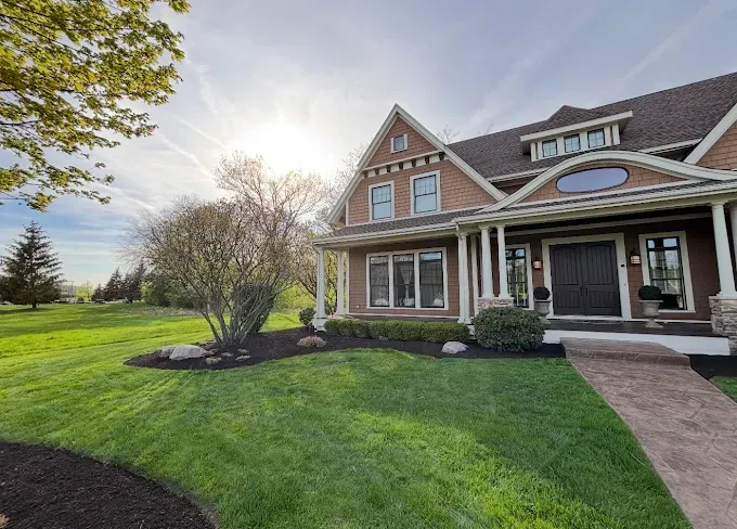 House with a brown exterior, green lawn, and blue sky. Sun shines brightly.