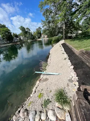 Lakefront with a gravel shoreline, small plants, and a diving board under a bright blue sky.