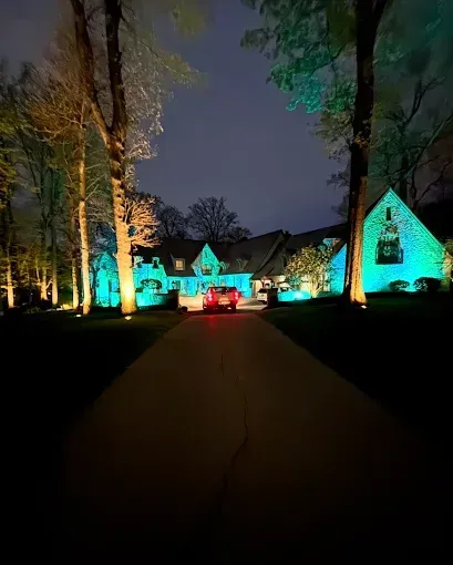 A car drives up a lit driveway to a house illuminated with green and blue lights at night.