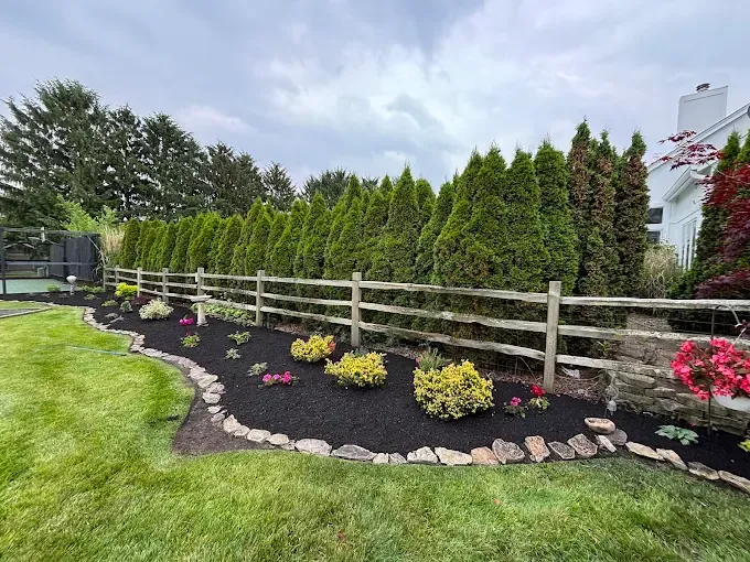 A landscaped garden with a split-rail fence, rows of green trees, and colorful flowers.