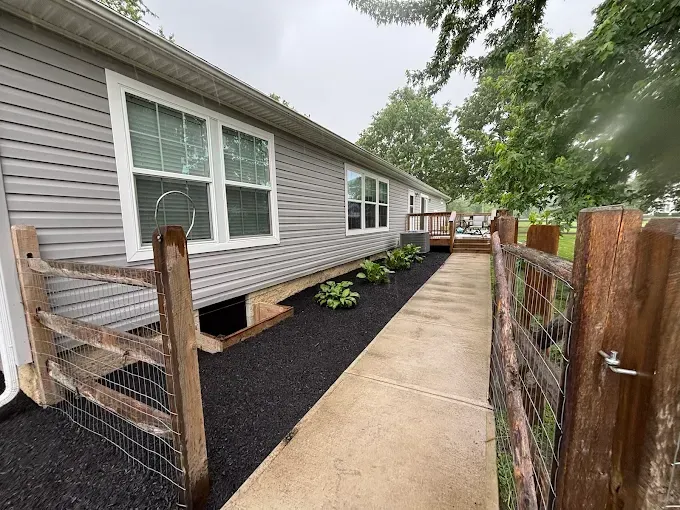 Exterior view of a gray house with a concrete walkway, mulch, and a wooden fence.