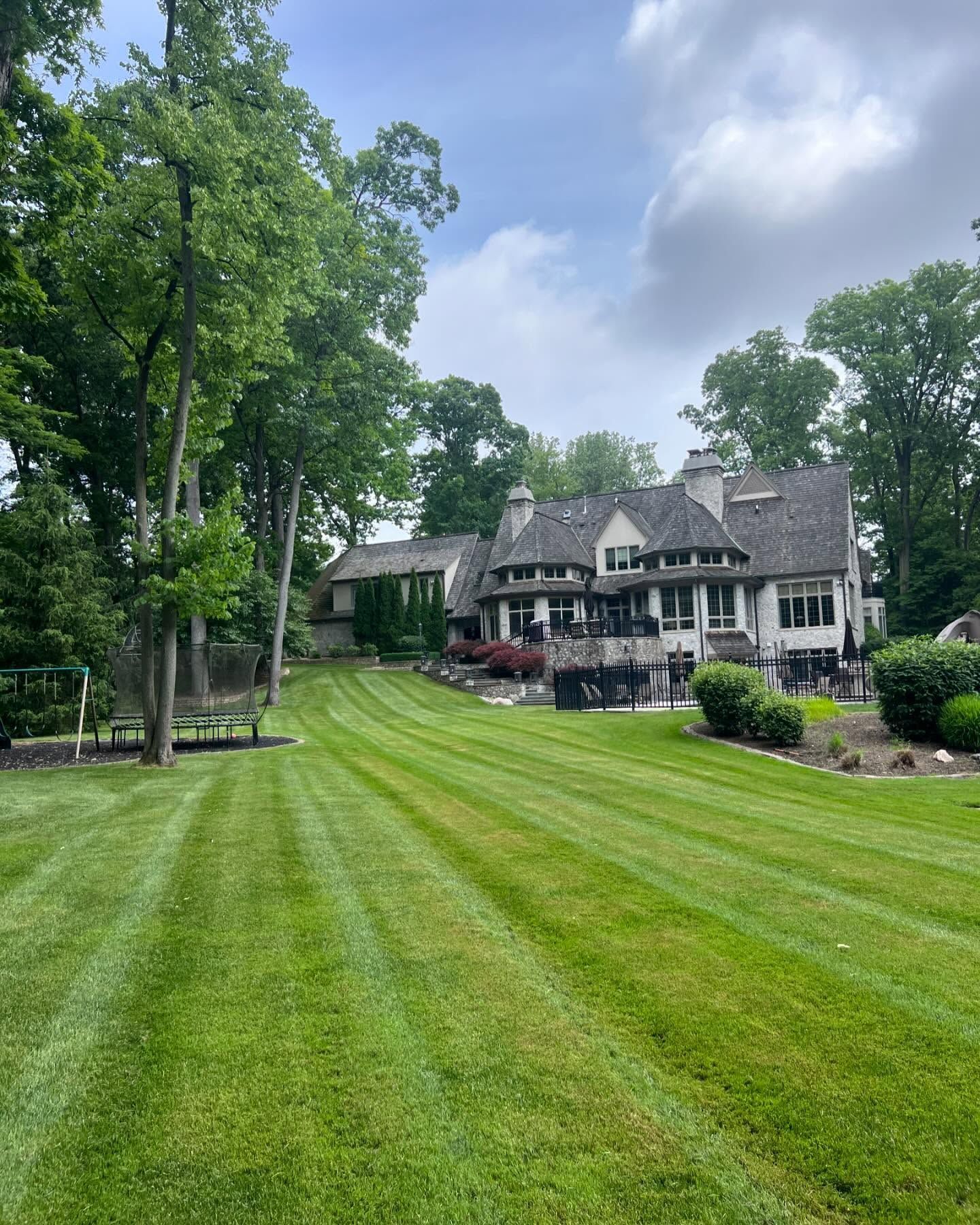 Well-manicured lawn leading to a large house with a gray roof, surrounded by trees on a cloudy day.