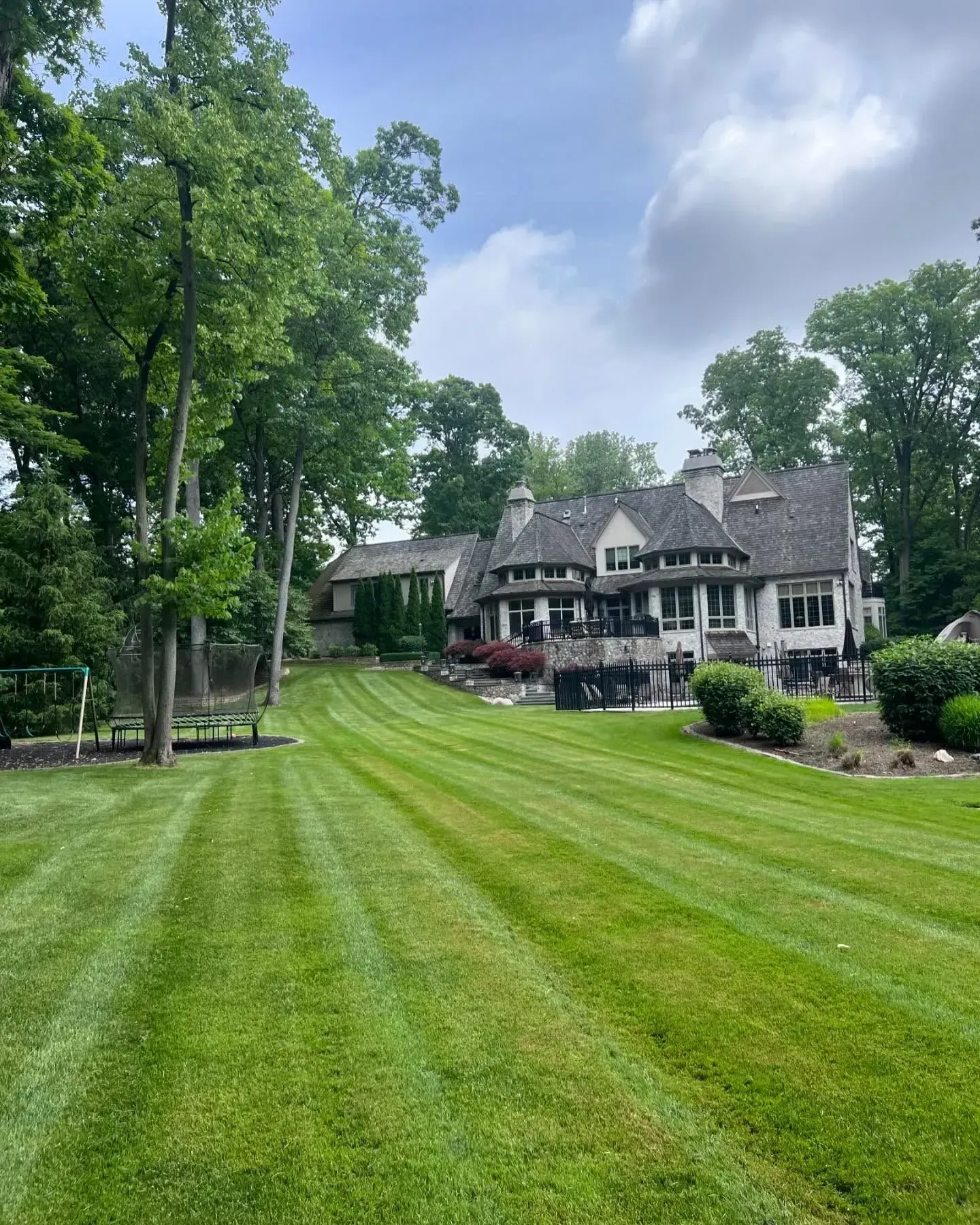 Large lawn leading to a Tudor-style house with trees and shrubs under a partly cloudy sky