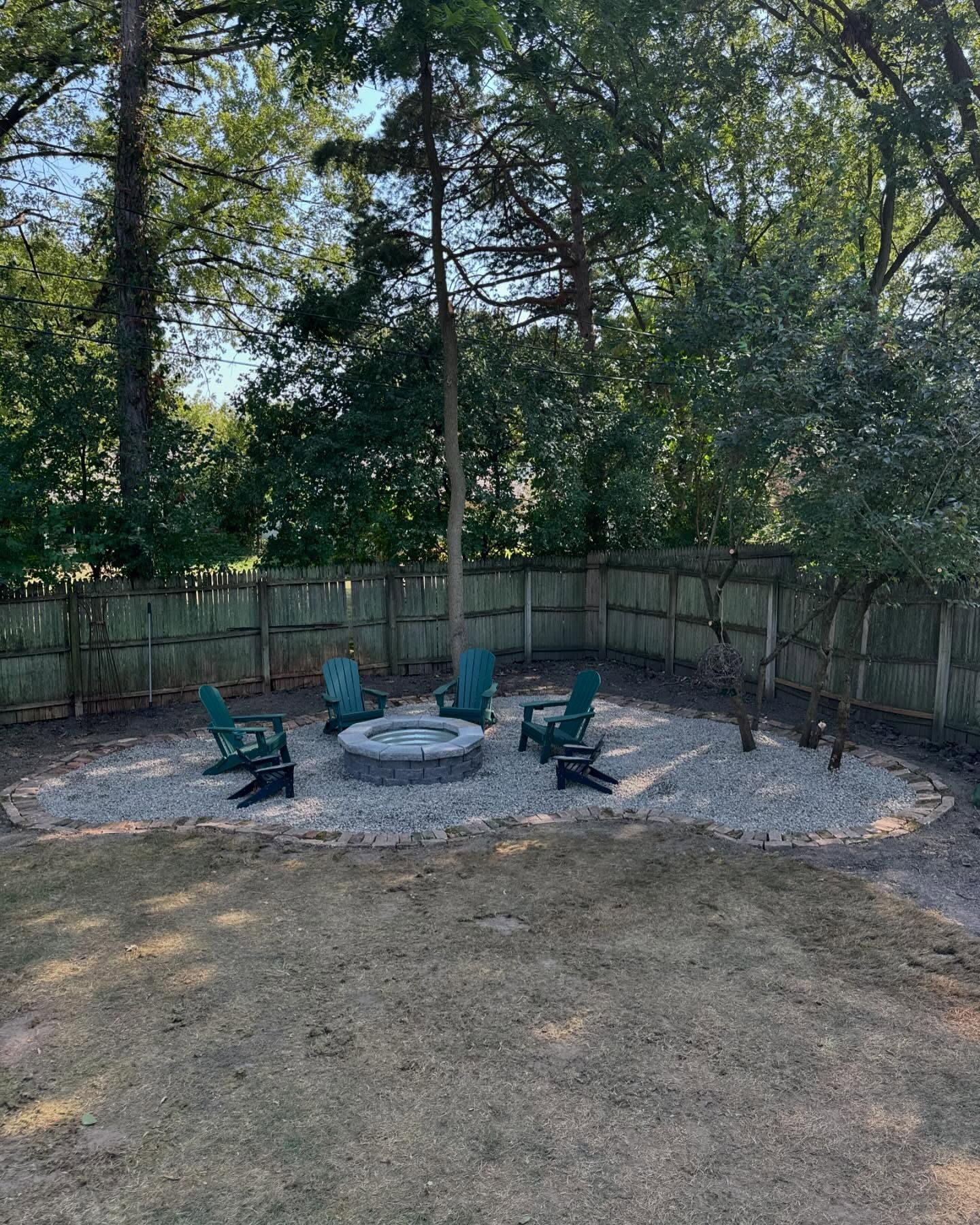 A backyard fire pit area with Adirondack chairs on gravel, surrounded by a wooden fence and trees.