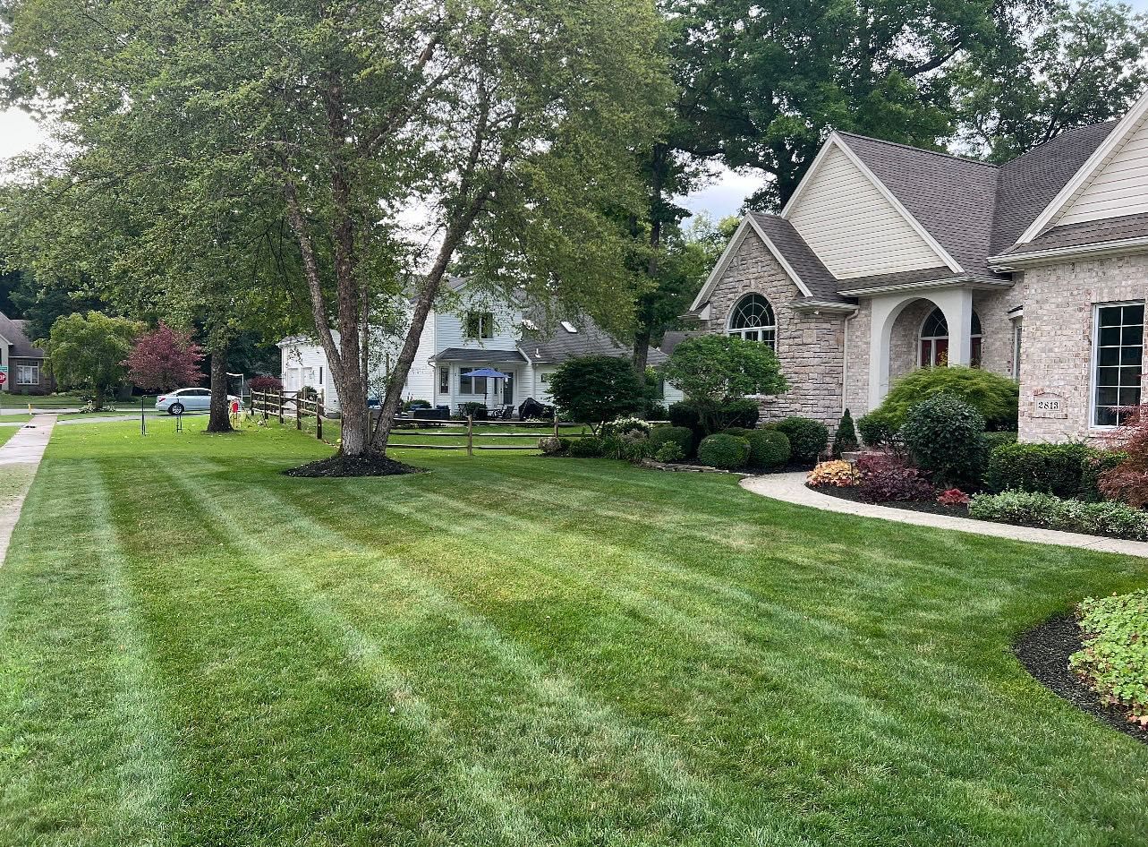 Lush green lawn mowed with stripes in front of a house with beige brick exterior and a tree.
