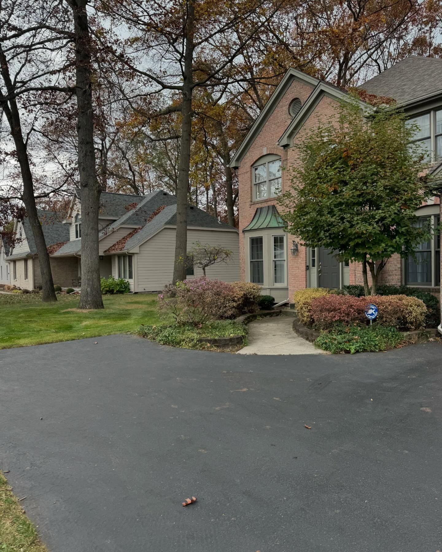 Brick house with black driveway and lush landscaping under a cloudy sky.