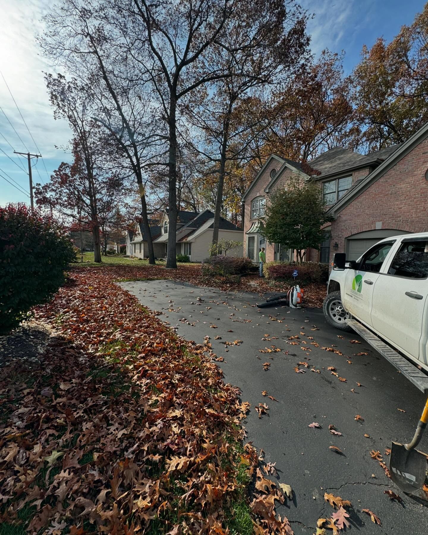 Leaves piled on a driveway next to a brick house and a white pickup truck on a sunny autumn day.