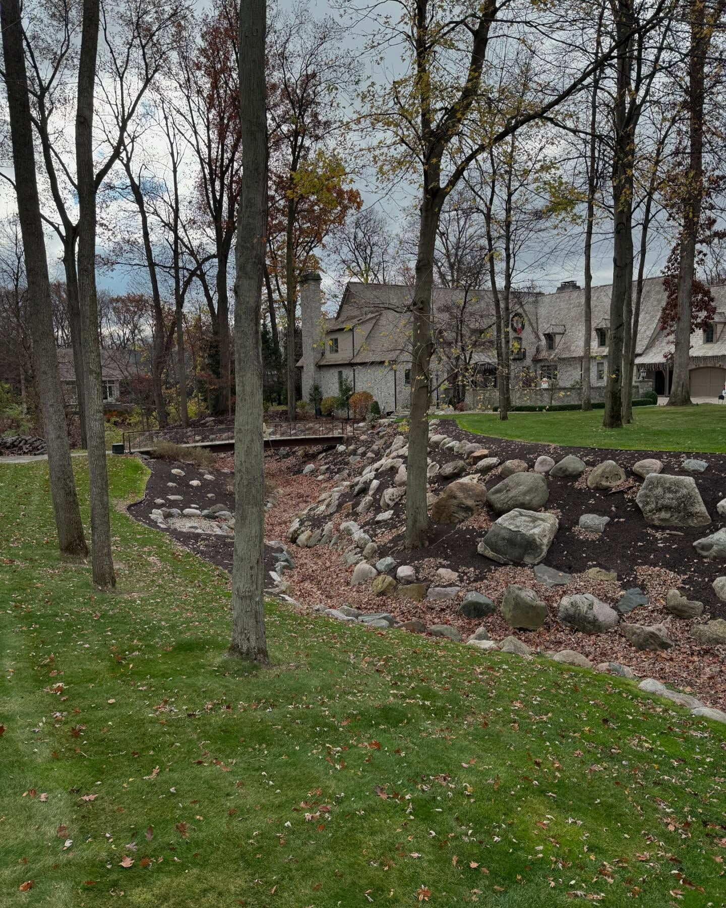 Lush green lawn with bare trees, a rocky stream bed, and a large house in the background under a cloudy sky.