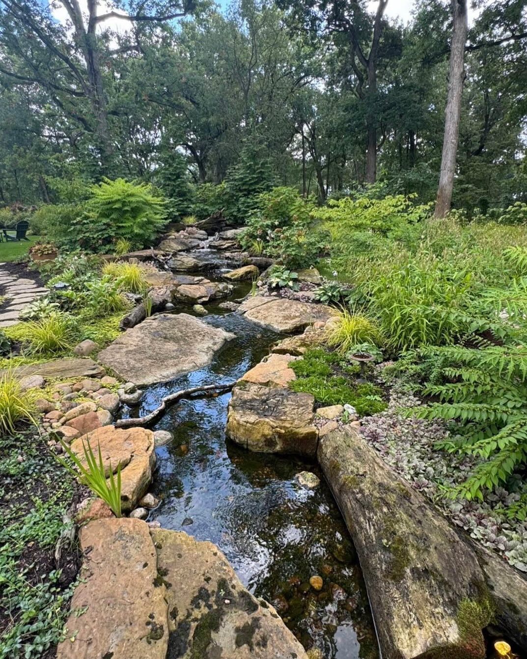 A stream flows through a lush garden with large rocks and green vegetation, trees in the background.