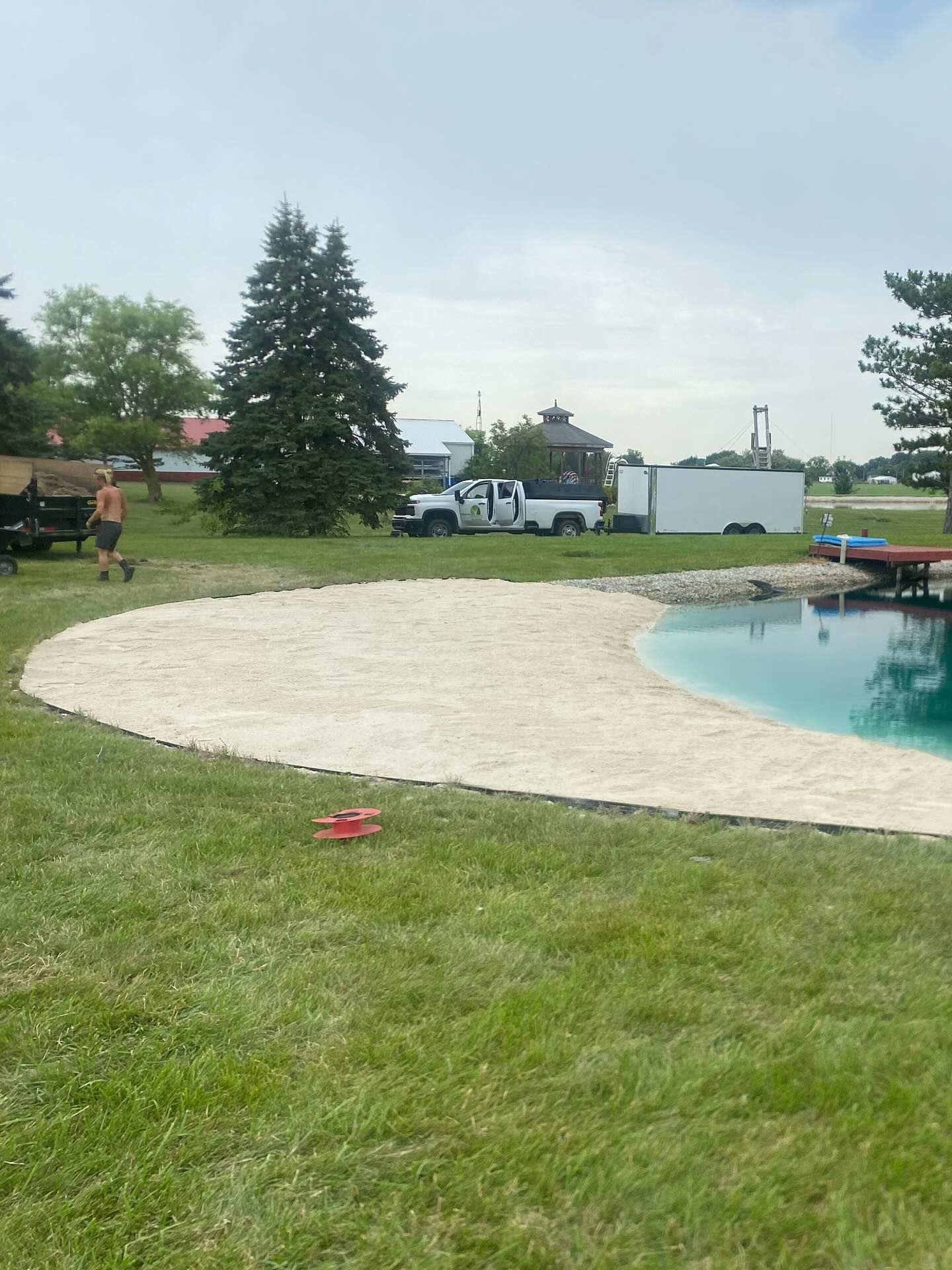 A yard with a beach area next to a pool. A person walks a dog on the grass.