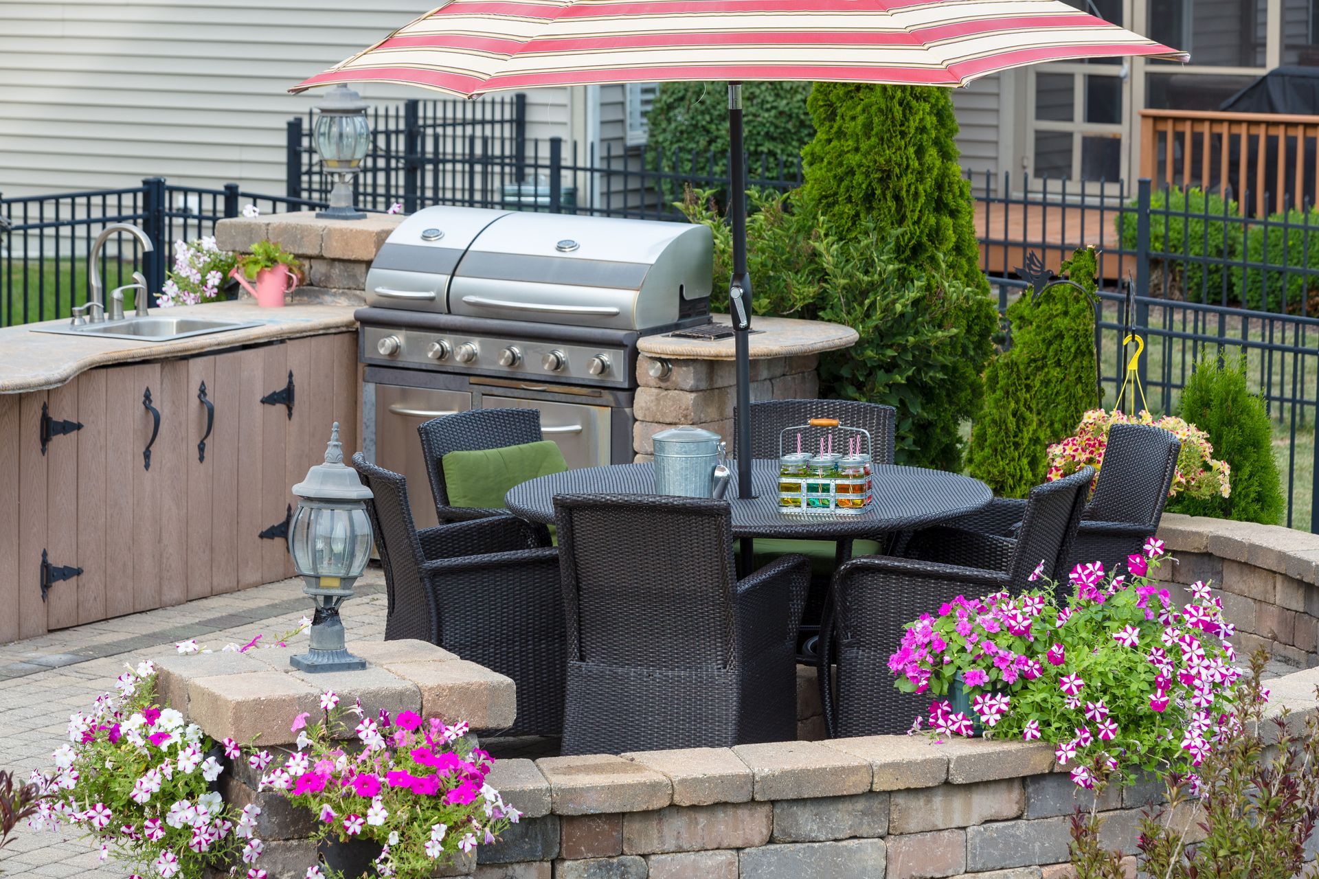 An outdoor kitchen area with a built-in grill, patio table with chairs under a striped umbrella, and floral landscaping.