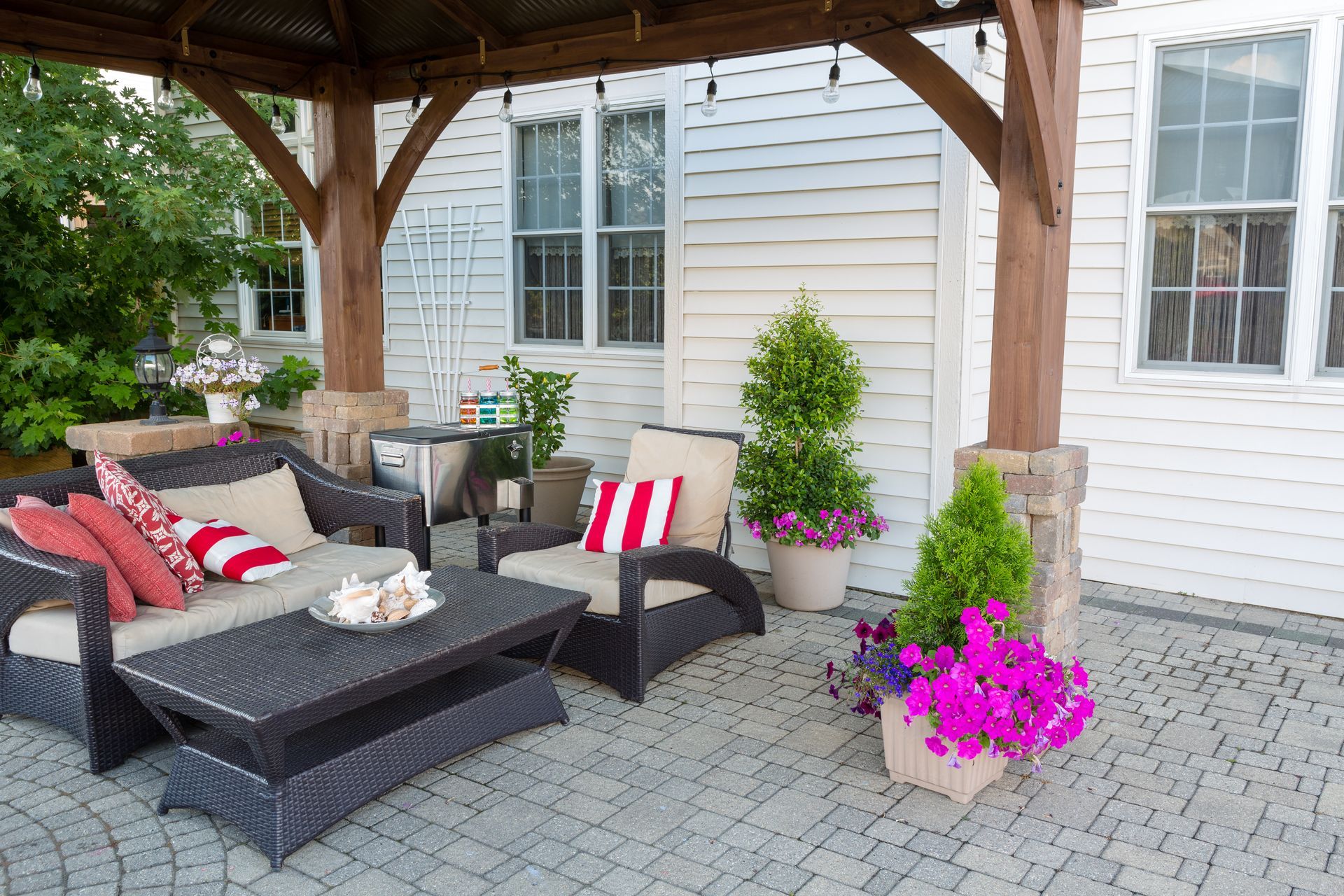 A patio with a dark wicker sofa and armchair, a coffee table, and potted plants under a wooden pergola against a house.