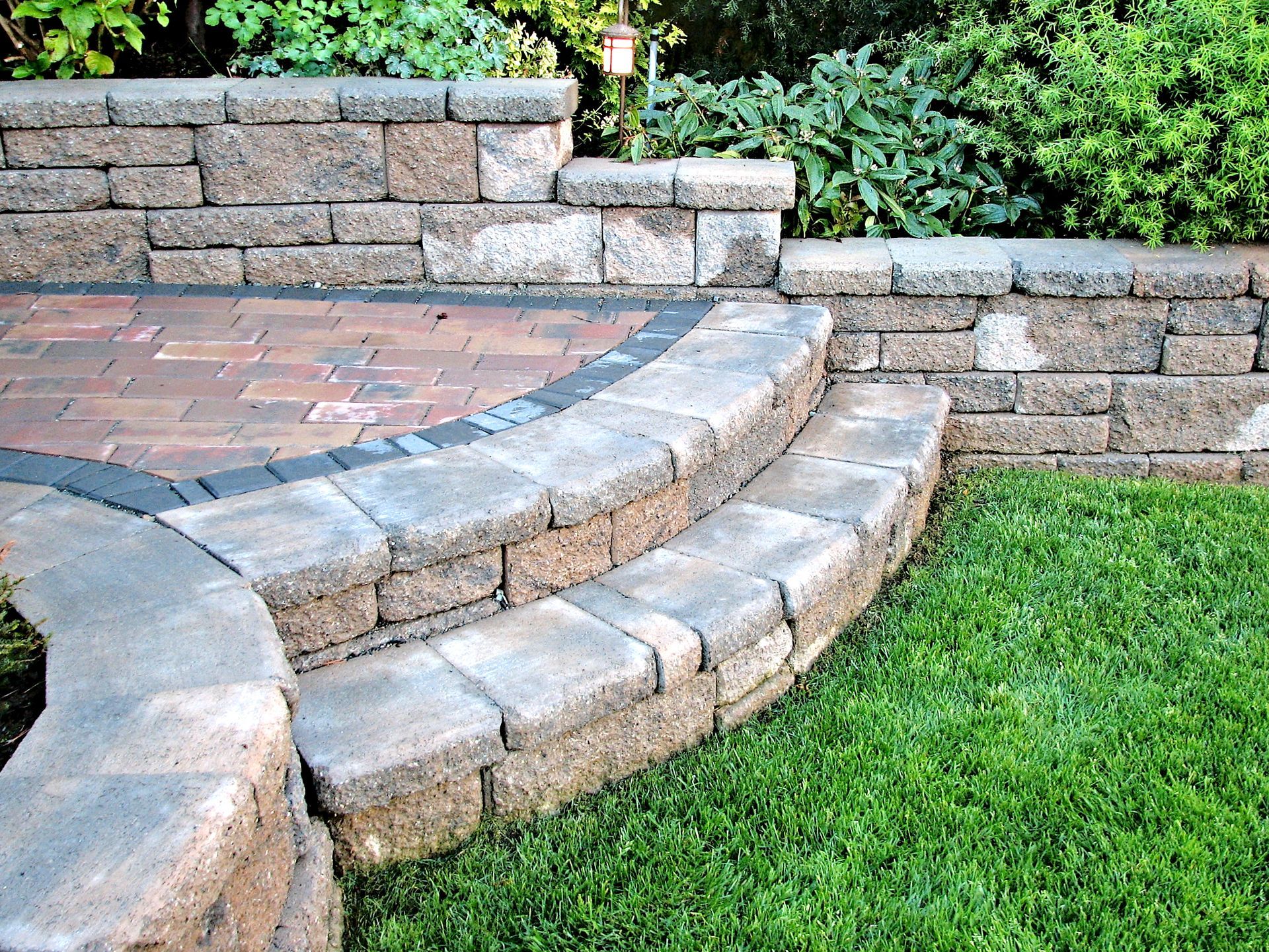 Stone steps lead up to a brick patio, surrounded by a retaining wall, and green grass.
