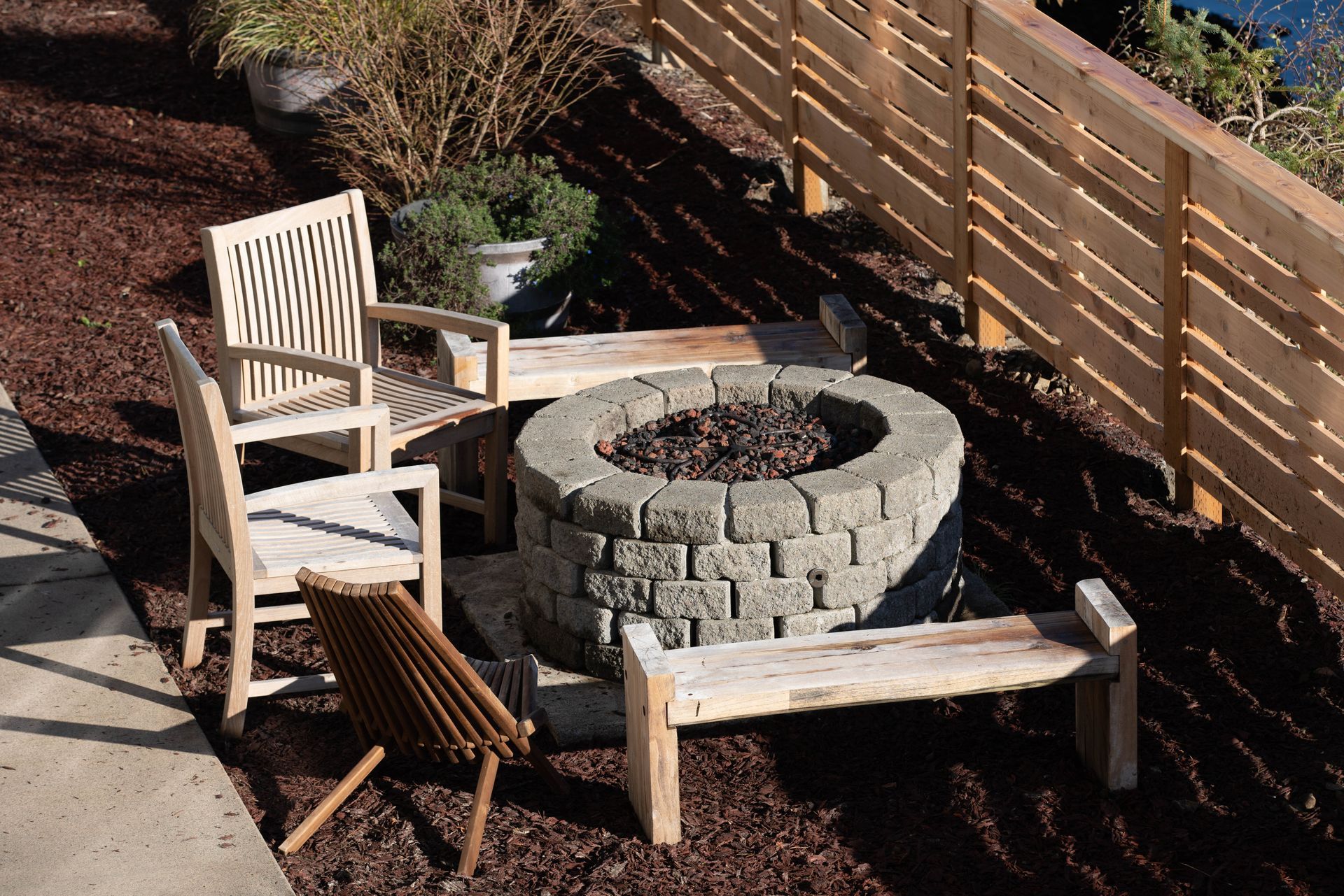 A stone fire pit surrounded by wood chairs and a bench on a mulch patio next to a wooden fence.