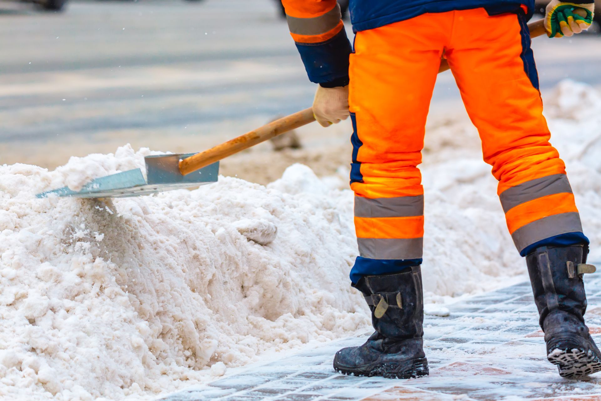 A worker in orange and navy high-visibility clothing clears a pile of snow from a walkway using a shovel.