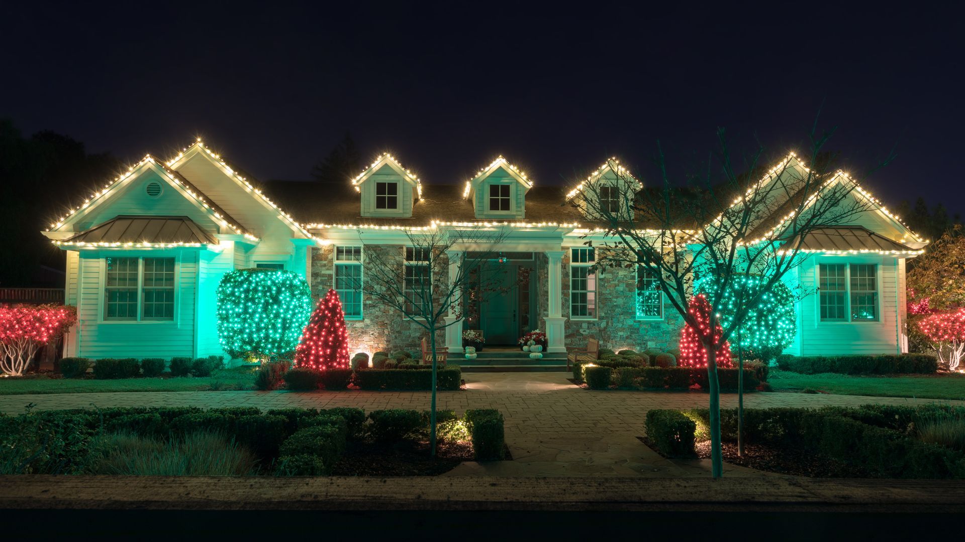 A one-story house at night, decorated with white roofline lights, green wall wash lighting, and two small red light trees.