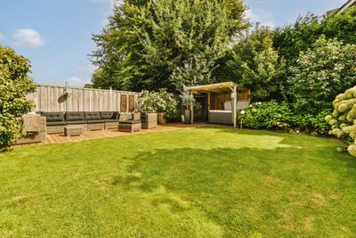 A sunny backyard featuring a lush green lawn, a wooden patio with modular seating, and a covered lounge area.