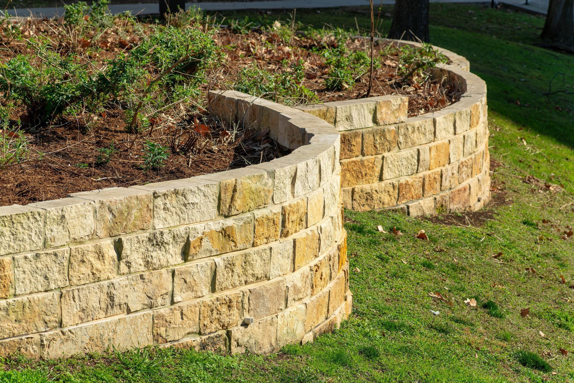 A curved, two-tier garden retaining wall made of stacked tan-colored, textured stone blocks, set in a grassy yard.