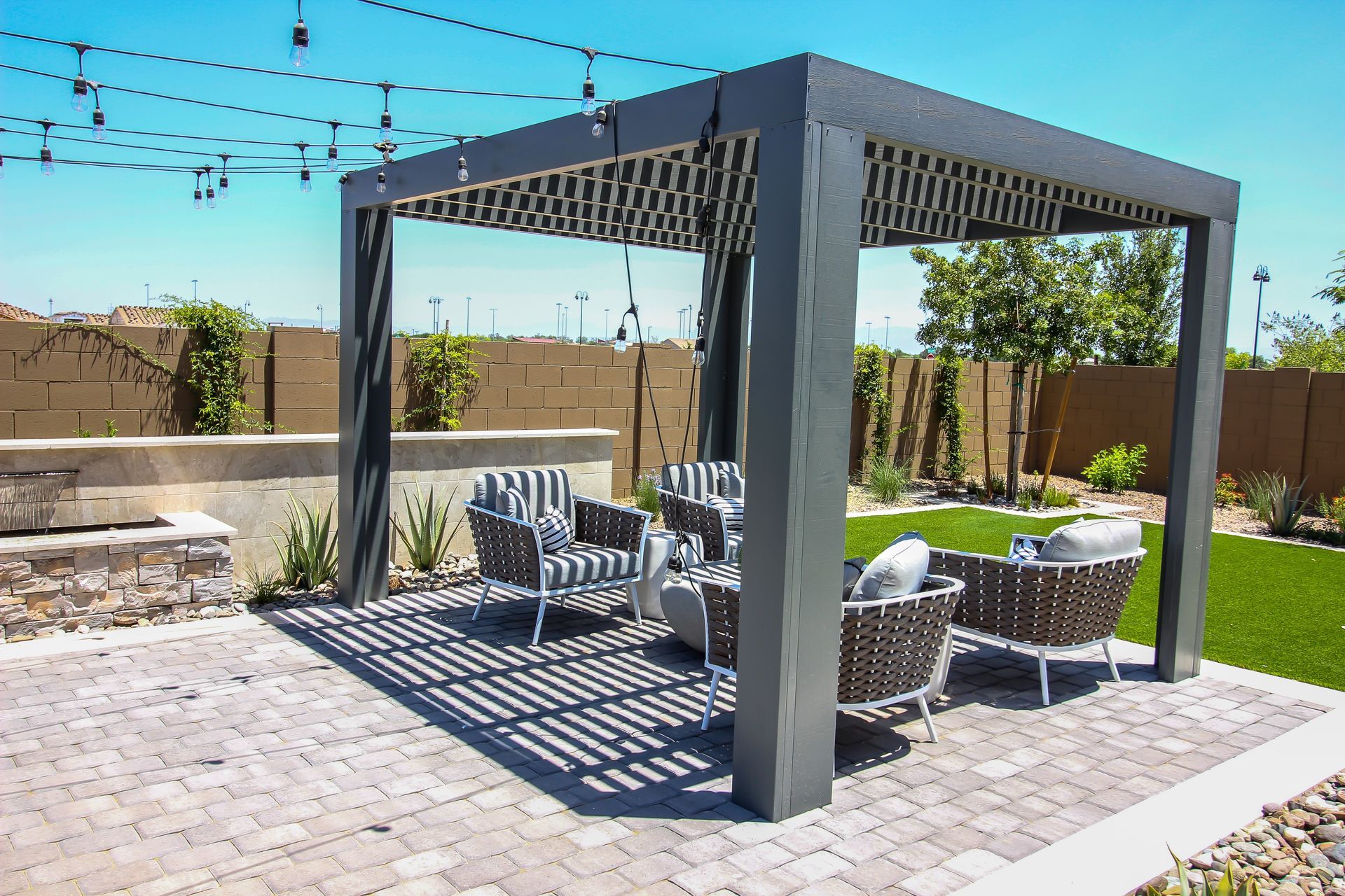 A modern gray pergola on a stone patio with wicker patio furniture, set in a sunny backyard with artificial grass.