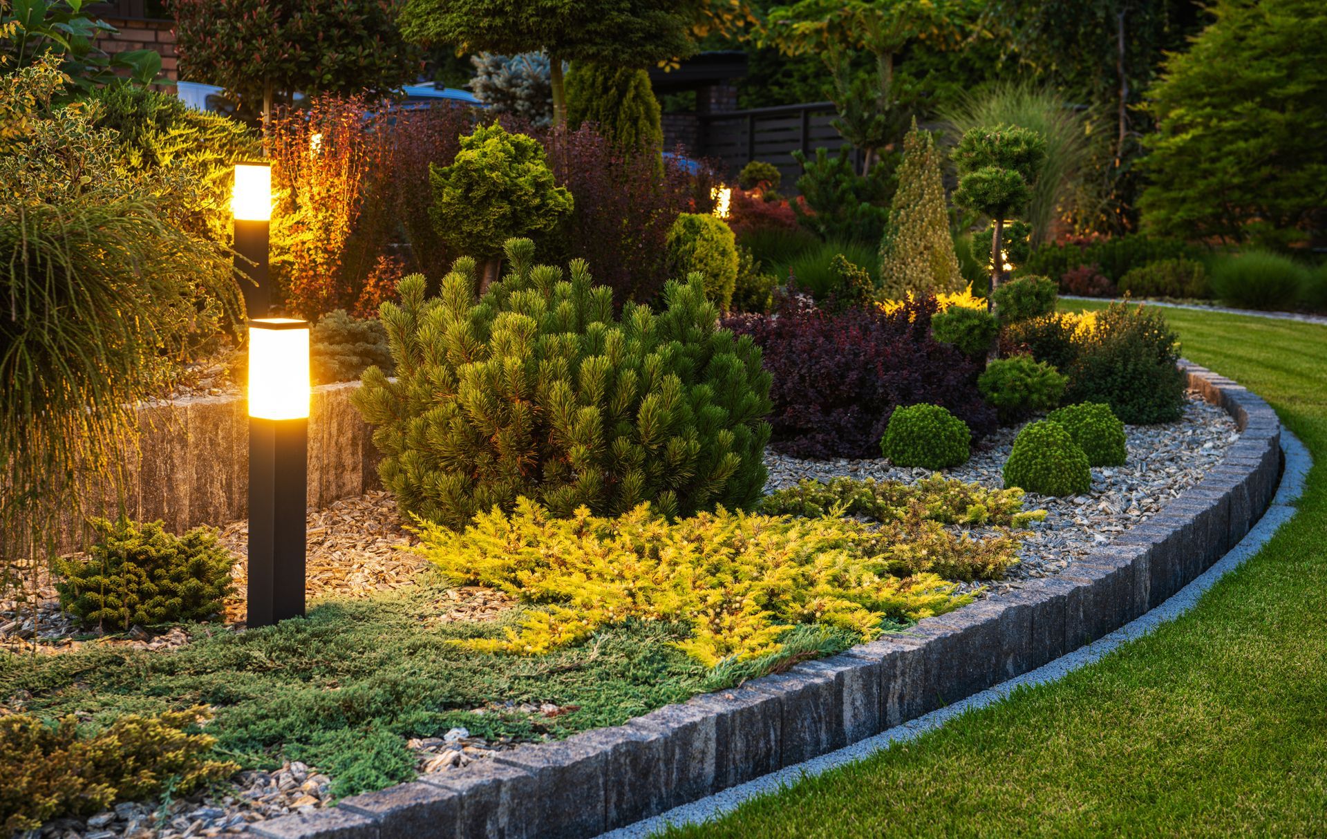 A landscaped garden bed at twilight with illuminated path lights, ornamental shrubs, gravel, and a stone-edged lawn.