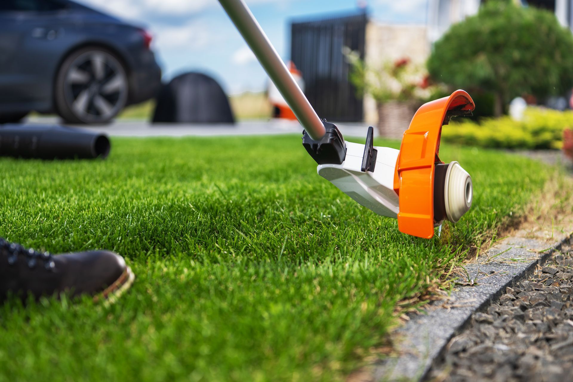 A person uses an orange and white string trimmer to edge a green lawn next to a gravel path and a parked car.