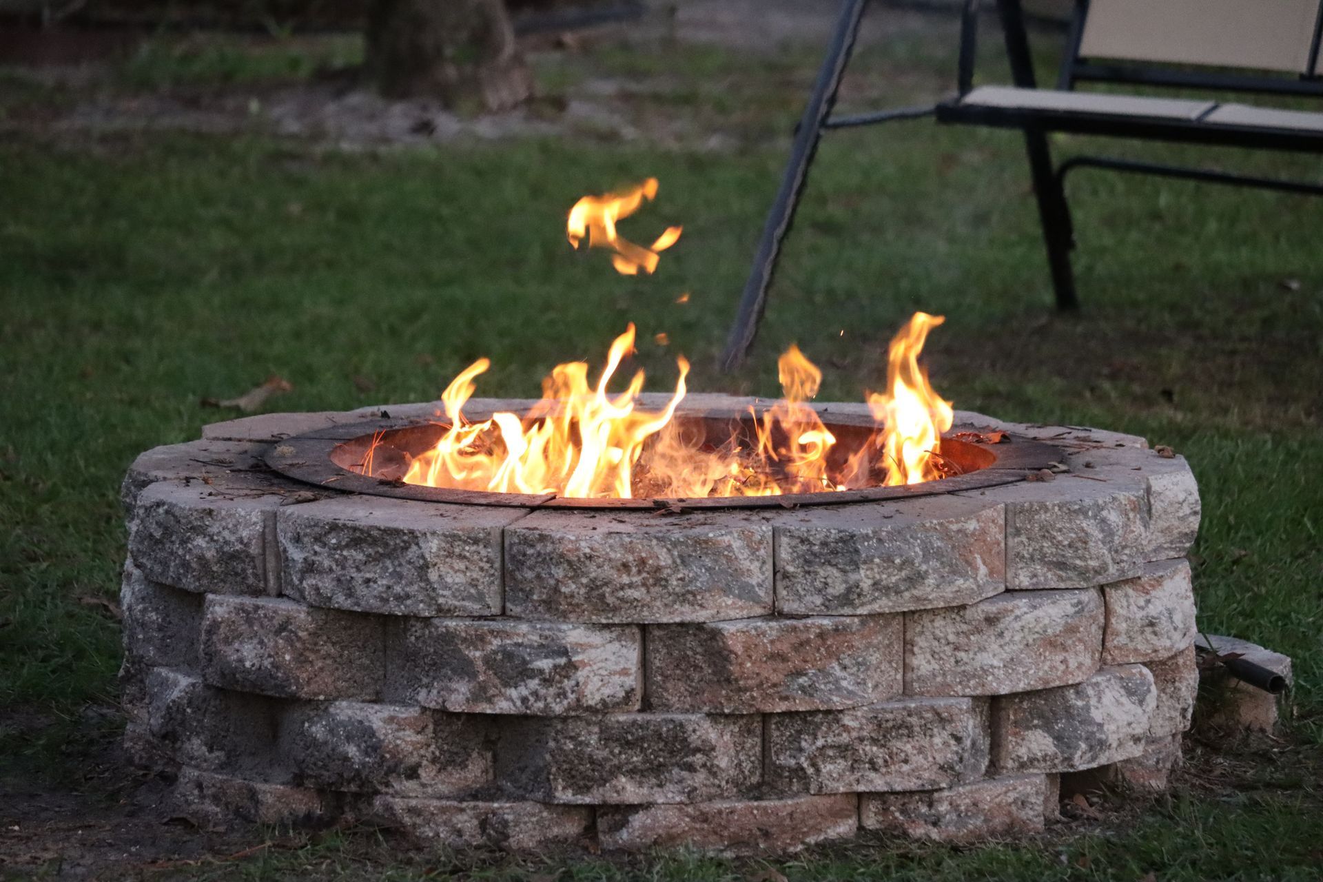 A circular stone fire pit burning with orange flames, set in a grassy backyard with a bench in the background.