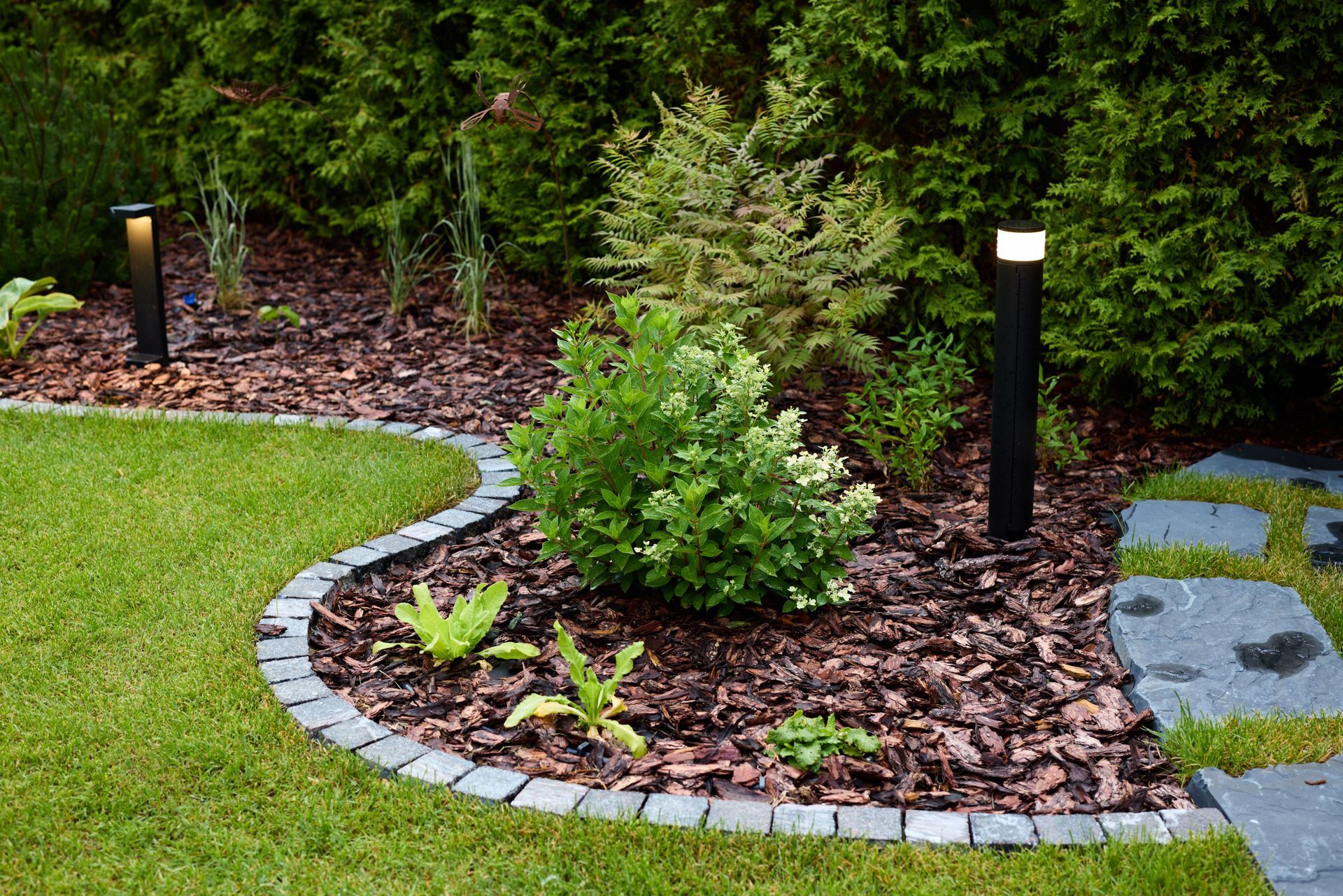 Curved stone-edged garden bed with shrubs, mulch, and black path lights beside a lawn and stepping stones