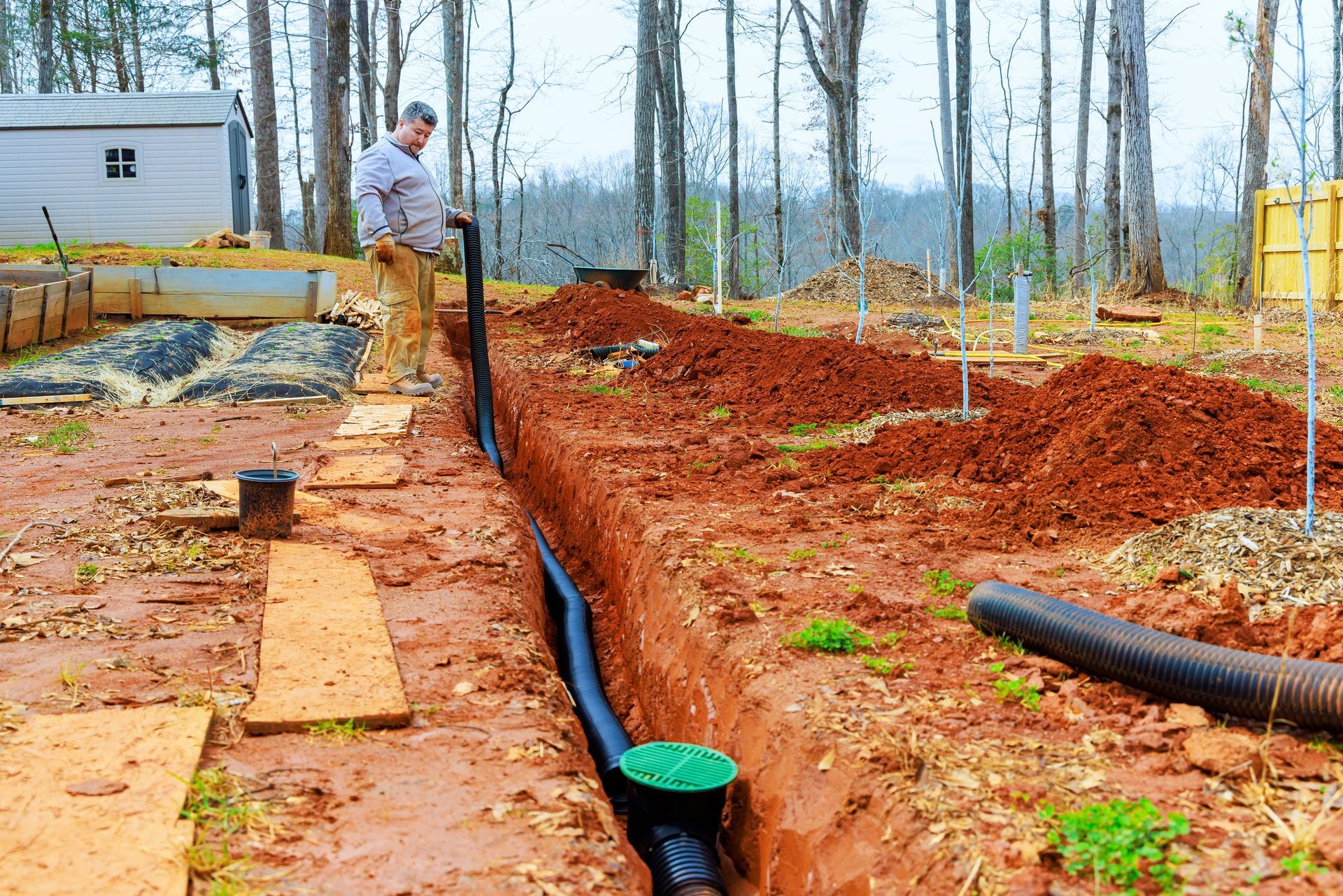 A person in a trench installing black drainage pipe with a green catch basin in a muddy, wooded backyard setting.