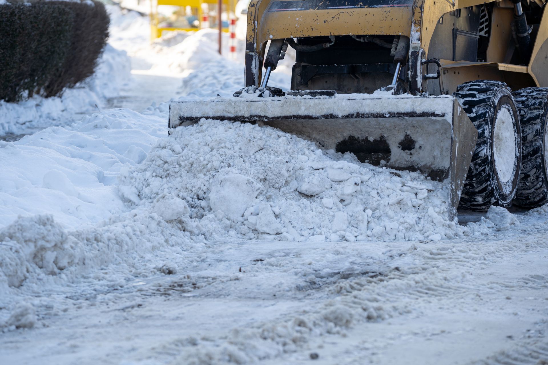A yellow skid steer loader pushes a large pile of snow across a snow-covered parking lot.