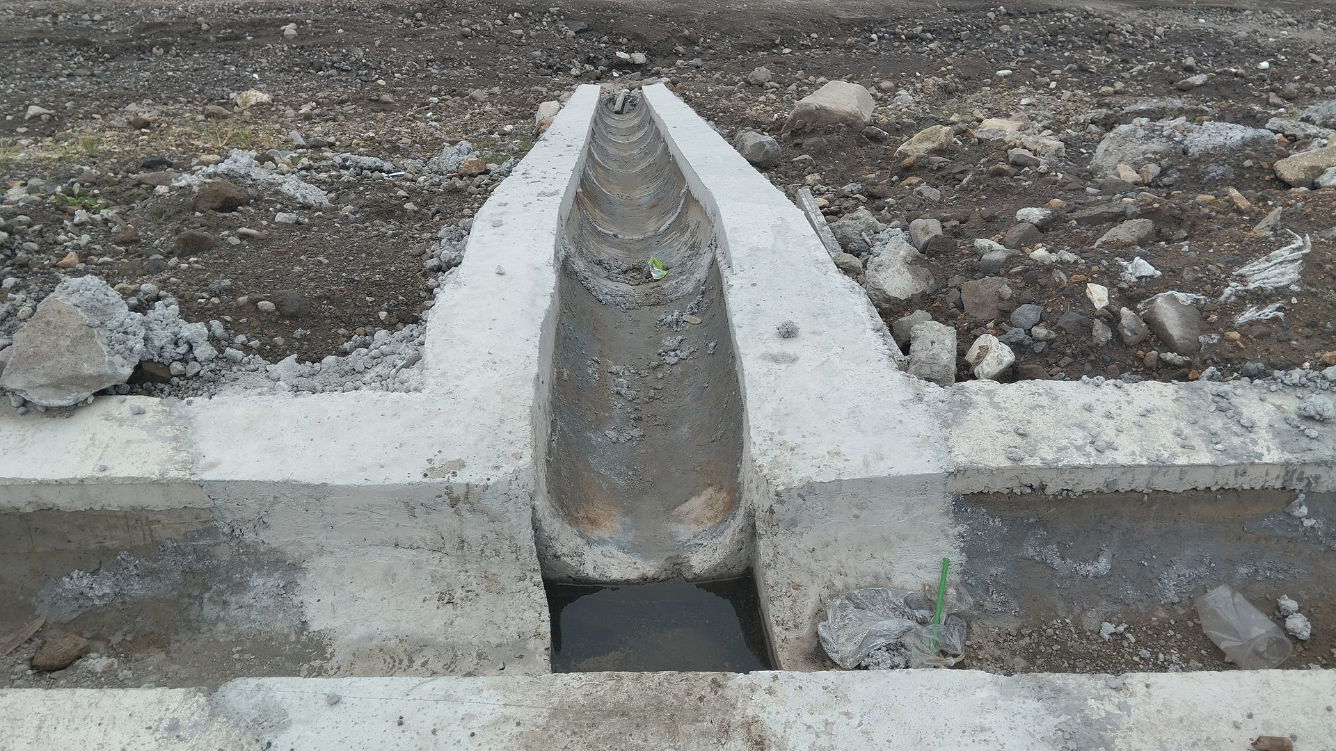 A concrete drainage channel intersection on dirt ground, with a curved center trough leading into a rectangular opening.
