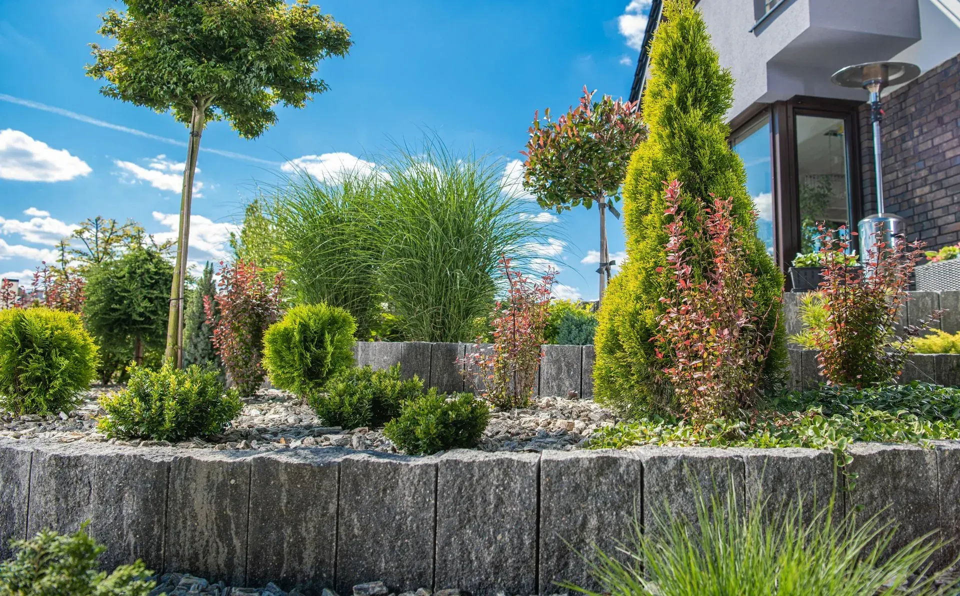 Landscaped garden with shrubs, flowers, and a small tree beside a modern house under a blue sky