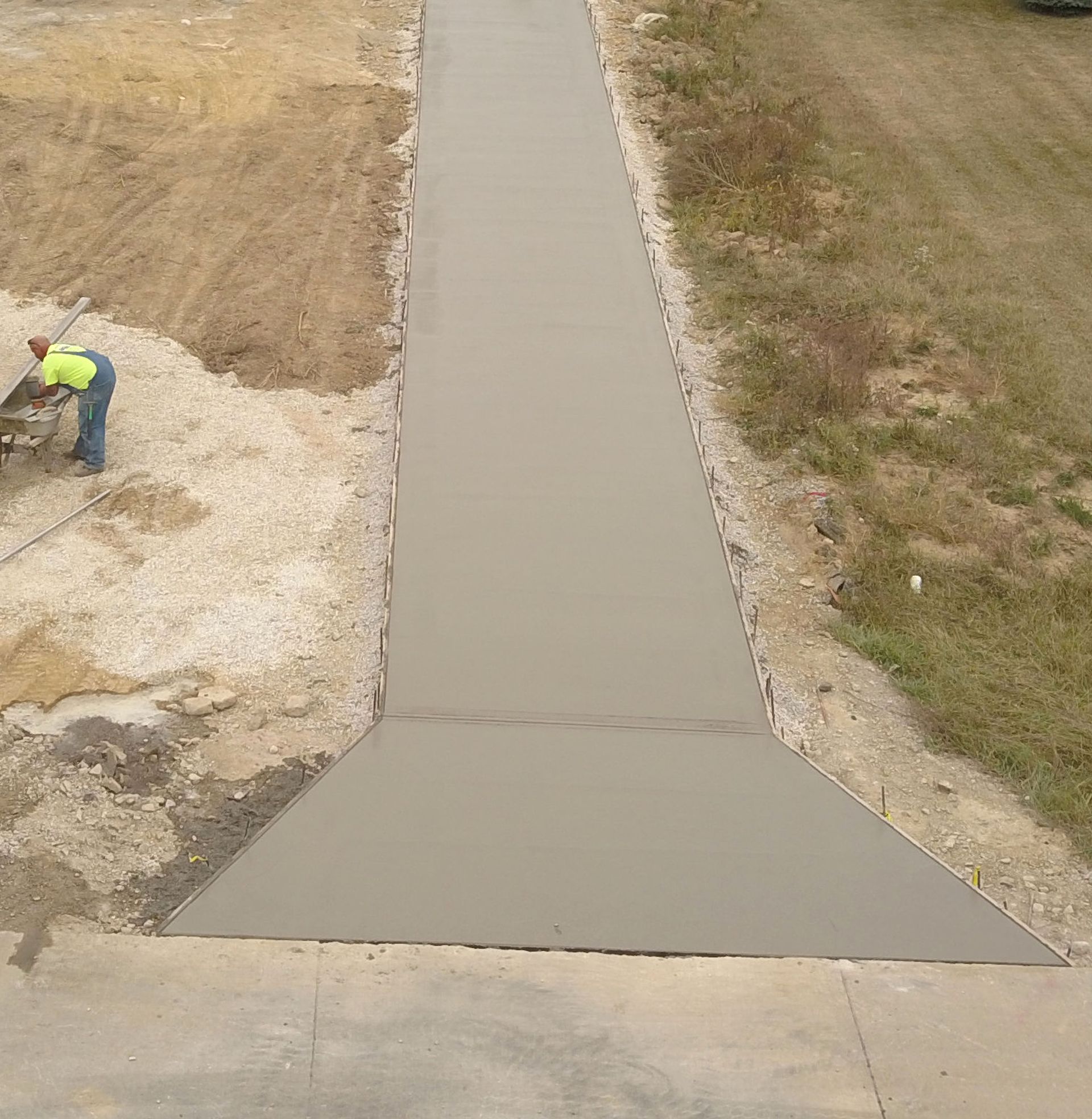 A person in a high-visibility vest stands next to a freshly poured concrete walkway that widens at the bottom.