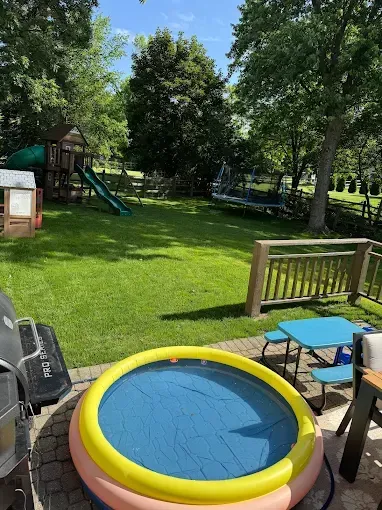 Backyard scene: Inflatable pool, green lawn, play structure, trees, small blue table.