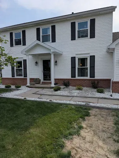 White two-story house with black shutters, red brick base, and white gravel landscaping under a blue sky.