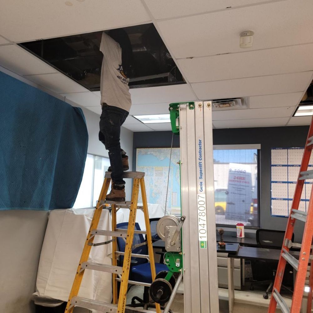 Person on ladder reaching into ceiling opening; lift and ladder present in office.