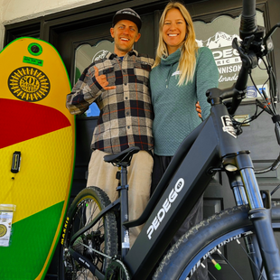 Two people pose with e-bikes beside a colorful surfboard display at a shop entrance