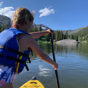 Paddleboarder in blue life vest paddling on a calm mountain lake under a clear sky