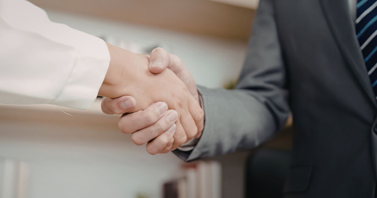 A business professional in a grey suit shakes hands with someone in a white long-sleeved shirt.