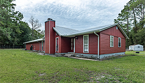 Red-sided house with a metal roof and a brick chimney on a green lawn with trees in the background.